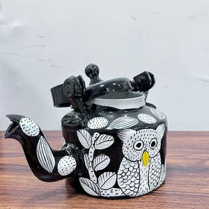 May include: Black metal tea kettle with a white owl and floral design. The kettle has a black handle and a black lid.