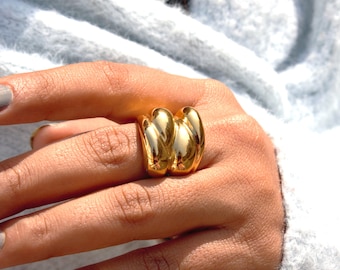 Può includere: Un anello dorato dal design unico, caratterizzato da tre fasce curve collegate. L'anello è mostrato su una mano, con un maglione azzurro chiaro sullo sfondo. La superficie lucida dell'anello riflette la luce, evidenziando la sua forma liscia e arrotondata.