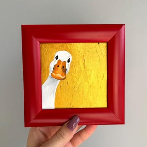Op de afbeelding: Een vierkant schilderij van een witte gans met een oranje snavel, geplaatst aan de linkerkant tegen een getextureerde metallic gouden achtergrond. Het kunstwerk is gevat in een dikke, glanzende rode lijst met afgeschuinde randen.