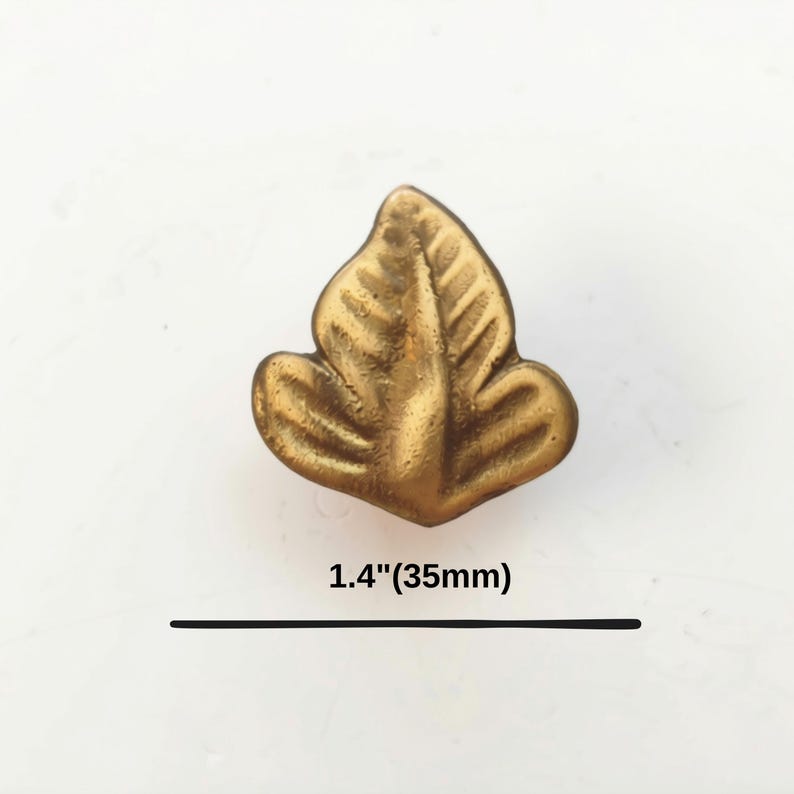 K&ouml;nnte beinhalten: Ein messingfarbener Metallknopf in Form eines dreilappigen Blattes mit gepr&auml;gten Aderdetails. Der Knopf hat eine strukturierte Oberfl&auml;che und misst 35 mm in der Breite, wie durch den Text und die Linie unter dem Objekt angegeben.