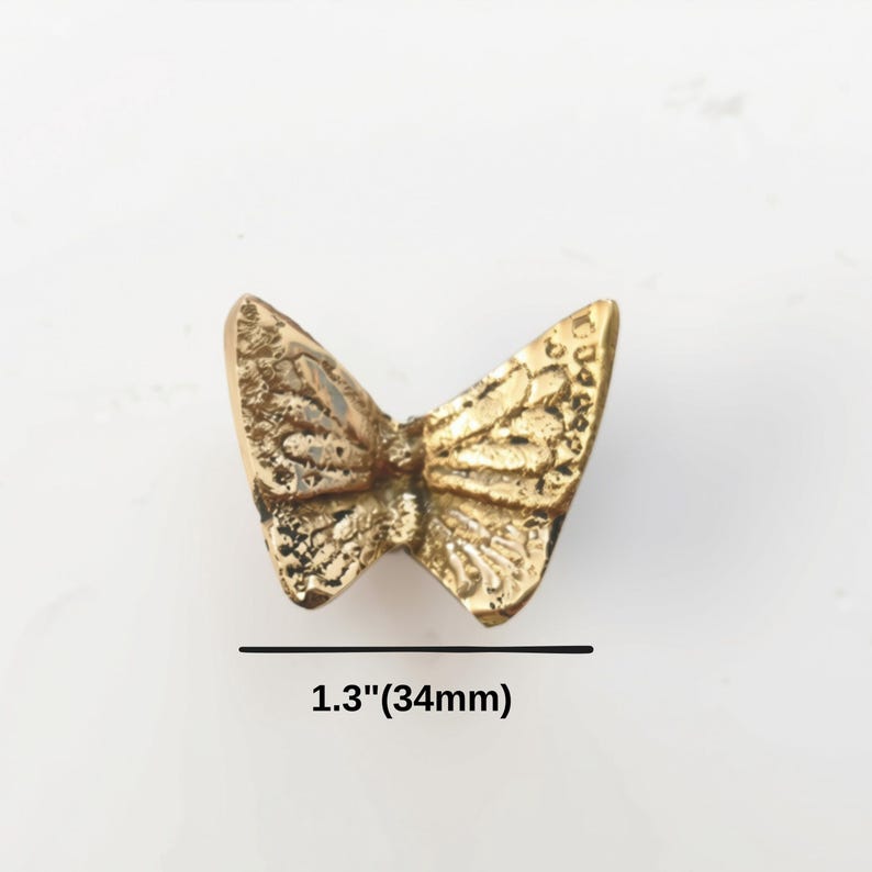 K&ouml;nnte beinhalten: Ein goldfarbener Metallknopf oder ein dekoratives St&uuml;ck in Schmetterlingsform mit einer texturierten Fl&uuml;geloberfl&auml;che. Das Objekt misst 34 mm in der Breite, wie durch den Text darunter angegeben. Das Design zeigt detaillierte, erhabene Fl&uuml;gelmuster.