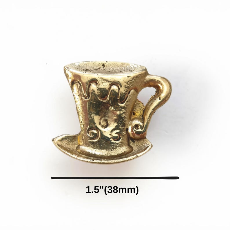 K&ouml;nnte beinhalten: Ein goldfarbener Teetassen-Anh&auml;nger aus Metall mit Untertasse, verziert mit gepr&auml;gten Schn&ouml;rkeln und einem geschwungenen Henkel. Eine schwarze Linie mit dem Text 1.5"(38mm) unter dem Objekt zeigt eine Breite von 38 mm an.