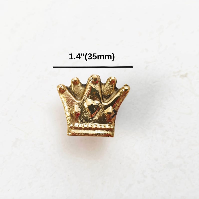 K&ouml;nnte beinhalten: Ein goldfarbener kronenf&ouml;rmiger Anh&auml;nger mit strukturierter Oberfl&auml;che und einem kleinen Herzdetail in der Mitte. &Uuml;ber dem Objekt steht der Text 1.4"(35mm), was eine Breite von 1,4 Zoll oder 35 Millimetern f&uuml;r dieses Dekorationselement angibt.