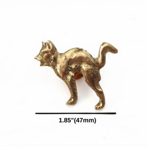 K&ouml;nnte beinhalten: Eine messingfarbene Metallkatzenfigur in einer Haltung mit gew&ouml;lbtem R&uuml;cken und erhobenem Schwanz. Die Katze ist mit offenem Maul und spitzen Ohren dargestellt. Das Objekt misst 47 mm in der L&auml;nge, wie durch den Text unter der Figur angegeben.