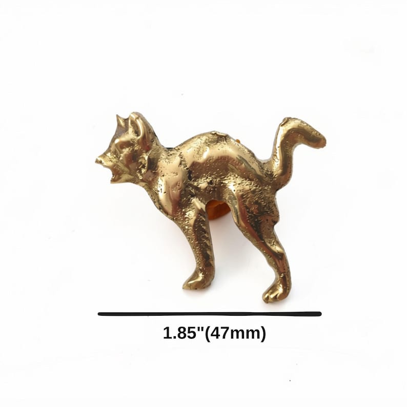 K&ouml;nnte beinhalten: Eine messingfarbene Metallkatzenfigur in einer Haltung mit gew&ouml;lbtem R&uuml;cken und erhobenem Schwanz. Die Katze ist mit offenem Maul und spitzen Ohren dargestellt. Das Objekt misst 47 mm in der L&auml;nge, wie durch den Text unter der Figur angegeben.