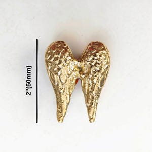 K&ouml;nnte beinhalten: Ein goldfarbenes Metallornament in Form von Engelsfl&uuml;geln mit einer texturierten, reflektierenden Oberfl&auml;che. Eine schwarze vertikale Linie neben den Fl&uuml;geln zeigt eine H&ouml;henmessung von 50 mm an. Die Fl&uuml;gel sind symmetrisch und nach unten gerichtet.