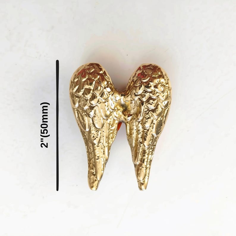 K&ouml;nnte beinhalten: Ein goldfarbenes Metallornament in Form von Engelsfl&uuml;geln mit einer texturierten, reflektierenden Oberfl&auml;che. Eine schwarze vertikale Linie neben den Fl&uuml;geln zeigt eine H&ouml;henmessung von 50 mm an. Die Fl&uuml;gel sind symmetrisch und nach unten gerichtet.