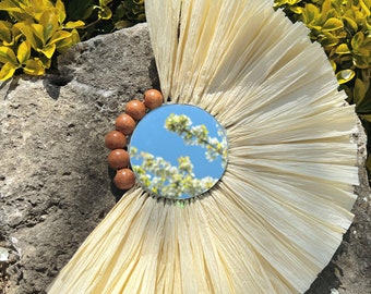 May include: A round mirror with a natural fiber frame and a brown wooden bead border. The mirror reflects a blue sky and white flowers.