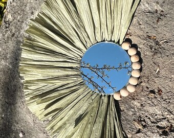 May include: A round mirror with a green paper mache frame and wooden beads around the edge. The mirror reflects a blue sky and a tree branch with white blossoms.