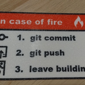 Może przedstawiać: Biały znak z czarnym tekstem i czerwonym paskiem u góry. Czerwony pasek zawiera tekst "W razie pożaru" i biały symbol płomienia. Znak zawiera trzy kroki: 1. git commit, 2. git push, 3. opuść budynek.