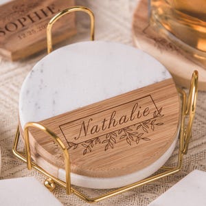 Könnte beinhalten: Ein Set personalisierter Untersetzer mit weißem Marmor- und Holzdesign. Die Untersetzer sind mit dem Namen "Nathalie" graviert und werden in einem goldenen Metallhalter aufbewahrt. Zusätzliche Untersetzer mit dem Namen "Sophie" sind ebenfalls sichtbar.