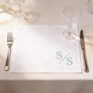 Puede incluir: Mantel individual rectangular blanco con un monograma bordado en azul claro con las letras "S/S". Un tenedor y un cuchillo plateados se colocan a cada lado del mantel individual. Un vaso de agua de cristal está colocado encima.