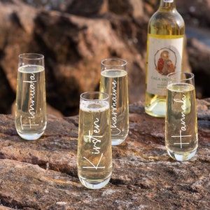 Op de afbeelding: Vier heldere glazen champagneflutes met gepersonaliseerde namen gegraveerd aan de zijkant. De namen zijn "Kirsten", "Charmaine", "Hamish" en "Fiona". De flutes staan op een houten oppervlak met een fles witte wijn op de achtergrond.