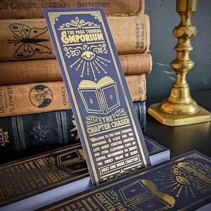 Può includere: Segnalibro blu navy con scritte dorate. Il segnalibro recita "The Page Turners Emporium" e "The Chapter Chaser". Il design include un occhio, un libro aperto e decorazioni. I libri sono sullo sfondo.