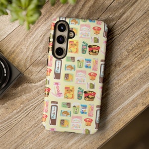 Puede incluir: Una funda de teléfono amarilla con un patrón de aperitivos y bebidas japonesas. La funda tiene un recorte de cámara negro y un borde blanco.