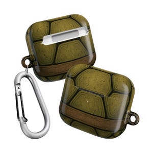 May include: Two olive green and brown AirPod cases with a turtle shell design. The cases have a silver carabiner clip attached. The cases are square with a rounded top and bottom. The cases are designed to protect the Airpods.