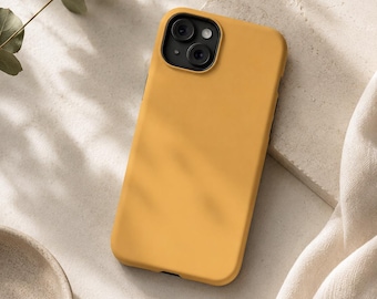 May include: Mustard yellow phone case with a black camera module. The case is lying on a textured surface, with a sprig of greenery and a small ceramic bowl in the background. The case has a smooth, matte finish.