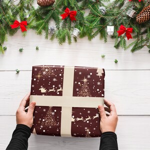 Puede incluir: Una persona est&aacute; envolviendo un regalo de Navidad con papel de regalo marr&oacute;n con un dise&ntilde;o de copo de nieve dorado y &aacute;rbol de Navidad. El papel tiene una cinta blanca atada alrededor.
