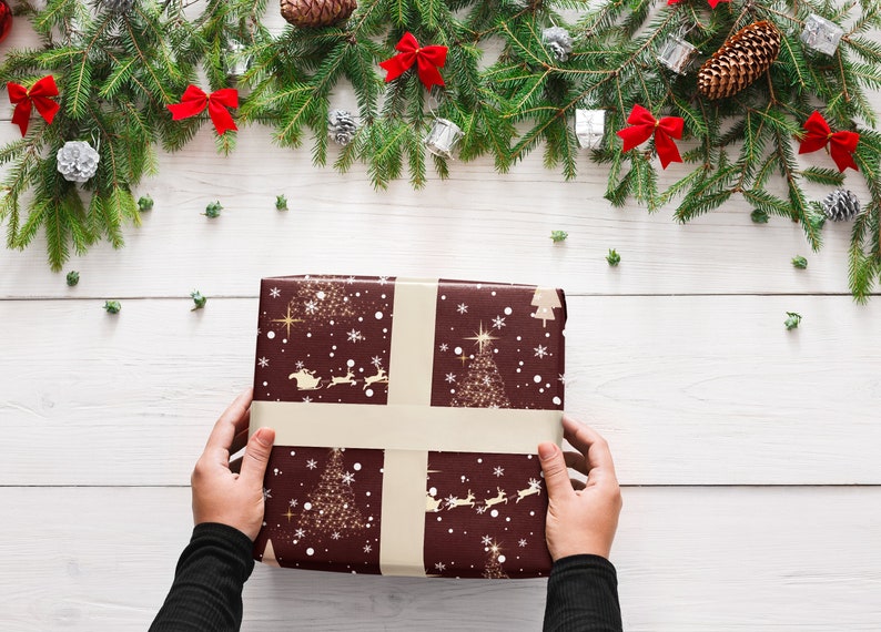 Puede incluir: Una persona est&aacute; envolviendo un regalo de Navidad con papel de regalo marr&oacute;n con un dise&ntilde;o de copo de nieve dorado y &aacute;rbol de Navidad. El papel tiene una cinta blanca atada alrededor.