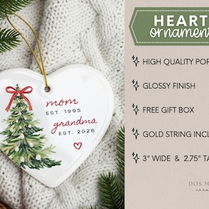 May include: A white heart-shaped porcelain ornament with a glossy finish, featuring a green Christmas tree illustration with a red bow, and the text "mom EST. 1995" and "grandma EST. 2026" with a small red heart. It includes a gold string and measures 3" wide by 2.75" tall.