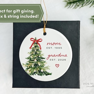 May include: A round white ceramic ornament featuring a watercolor illustration of a green Christmas tree topped with a red bow. The text reads mom EST. 1995 and grandma EST. 2026 with a small red heart below. A gold string is attached for hanging.