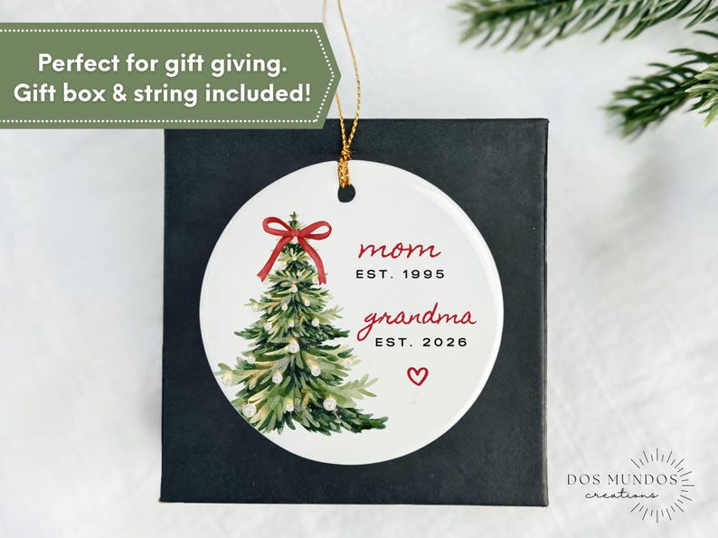 May include: A round white ceramic ornament featuring a watercolor illustration of a green Christmas tree topped with a red bow. The text reads mom EST. 1995 and grandma EST. 2026 with a small red heart below. A gold string is attached for hanging.