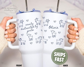 Puede incluir: Dos vasos blancos con asas y tapas transparentes, cada uno con una pajita de metal. El diseño incluye dibujos de líneas azul oscuro de cabras pequeñas y corazones. Un vaso muestra el texto "easily distracted by goats" y el otro "Olivia".