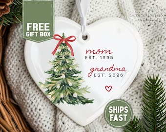 Puede incluir: Un adorno de cerámica blanco en forma de corazón con un árbol de Navidad en acuarela y un lazo rojo en la parte superior. El texto dice mom EST. 1995 y grandma EST. 2026, junto a un pequeño corazón rojo. Incluye una cinta blanca para colgar.