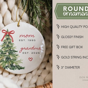 May include: A round white porcelain ornament with a 3-inch diameter, featuring a watercolor Christmas tree with a red bow, the text "mom EST. 1995" and "grandma EST. 2026" in red, a small red heart, and a gold string for hanging.