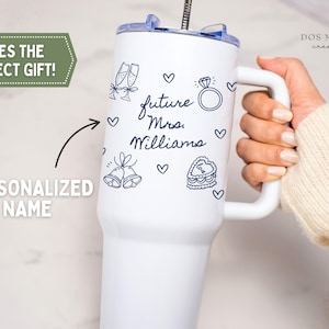 Puede incluir: Un vaso blanco de acero inoxidable con asa y tapa transparente que presenta ilustraciones de l&iacute;neas azul oscuro de copas de champ&aacute;n, un anillo de diamantes, campanas de boda y un pastel en forma de coraz&oacute;n con el texto future Mrs. Williams.