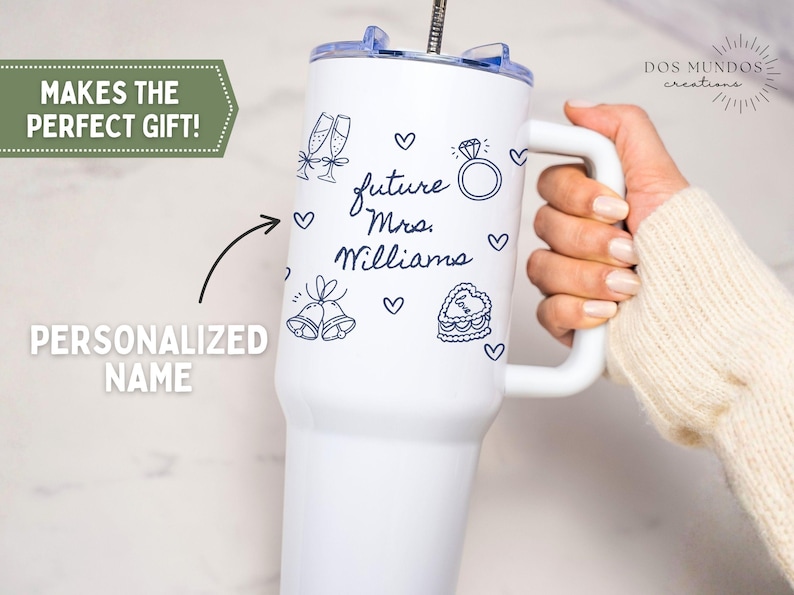 Puede incluir: Un vaso blanco de acero inoxidable con asa y tapa transparente que presenta ilustraciones de l&iacute;neas azul oscuro de copas de champ&aacute;n, un anillo de diamantes, campanas de boda y un pastel en forma de coraz&oacute;n con el texto future Mrs. Williams.
