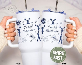 Puede incluir: Un vaso blanco de acero inoxidable con asa y tapa transparente que incluye una pajita de metal. El exterior está decorado con gráficos de porristas en azul oscuro, incluyendo pompones, megáfonos, lazos, estrellas y el texto Coach Madison.