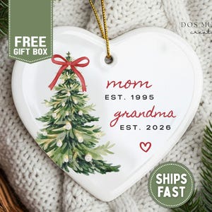 May include: A white heart-shaped ceramic ornament featuring a watercolor-style green Christmas tree with a red bow on top. The text reads mom EST. 1995 and grandma EST. 2026, with a small red heart below. A gold string is attached for hanging.