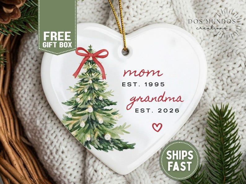 May include: A white heart-shaped ceramic ornament featuring a watercolor-style green Christmas tree with a red bow on top. The text reads mom EST. 1995 and grandma EST. 2026, with a small red heart below. A gold string is attached for hanging.