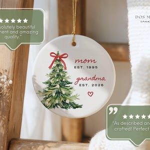 May include: A round white ceramic ornament featuring a watercolor illustration of a green Christmas tree topped with a red bow. The text reads mom EST. 1995 and grandma EST. 2026 with a small red heart below. It hangs from a gold string.