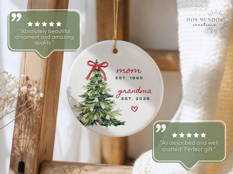 May include: A round white ceramic ornament featuring a watercolor illustration of a green Christmas tree topped with a red bow. The text reads mom EST. 1995 and grandma EST. 2026 with a small red heart below. It hangs from a gold string.
