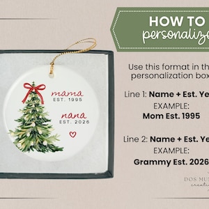 May include: A round white ceramic Christmas tree ornament with a gold hanging string, featuring a watercolor tree with a red bow, the text "mama EST. 1995" and "nana EST. 2026" with a red heart, presented in a black box with personalization instructions.