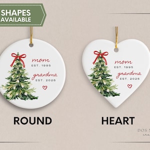 May include: Two white ceramic ornaments, one round and one heart-shaped, each featuring a watercolor Christmas tree with a red bow, the text mom EST. 1995, grandma EST. 2026, and a small red heart. Both ornaments hang from gold strings.