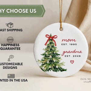 May include: A round white ceramic ornament hanging from a gold string. It features a watercolor green Christmas tree with a red bow on top. The text reads mom EST. 1995 and grandma EST. 2026 in red and black, with a small red heart below the text.