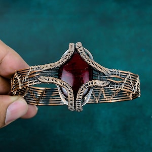 Pode incluir: Uma pulseira tipo bracelete com uma pedra preciosa oval de cor vermelho-escura ao centro, envolvida por um intrincado entrelaçamento de fio de cobre oxidado. O metal apresenta vários fios paralelos e bobinas apertadas em forma de gaiola.