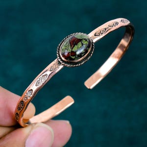 May include: A copper cuff bracelet featuring a central oval-shaped stone with mottled green and dark red patterns. The metal band is decorated with repeating geometric engravings and a twisted rope border surrounding the stone setting.