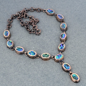Könnte beinhalten: Eine kupferfarbene Halskette mit ovalen Abalone-Muscheleinsätzen. Die schillernden Muscheln zeigen Farbtöne von Blau, Grün und Lila. Die Halskette hat eine Kette und einen Anhänger mit drei Abalone-Einsätzen.