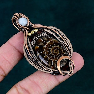 May include: A wire-wrapped pendant featuring a central oval ammonite fossil with a brown spiral pattern, encased in dark copper wire. The design includes a small white moonstone at the top and three tiny gold-toned beads near the fossil.