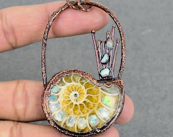 Puede incluir: Un colgante envuelto en alambre de cobre que presenta un amonite fosilizado con acentos de ópalo iridiscente. El colgante tiene un diseño delicado y orgánico con un grupo de piedras de ópalo en la parte superior.