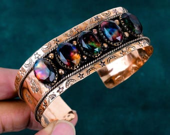 May include: A copper-toned cuff bracelet featuring five oval-shaped, faceted iridescent stones set in a row. The metal band is decorated with embossed star and crescent moon symbols and a twisted rope texture bordering the central stone setting.