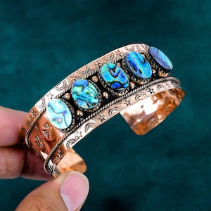 May include: A copper-toned cuff bracelet featuring five oval-shaped iridescent abalone shell inlays arranged in a row. The metal surface is embossed with repeating crescent moon and star patterns, bordered by thin twisted wire detailing.