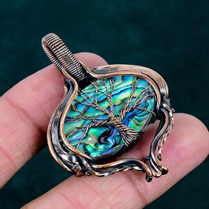Peut inclure: Un pendentif en fil métallique enveloppé avec une incrustation de coquille d'ormeau ovale irisée. La coquille présente des teintes vives de vert et de bleu. Un motif d'arbre en fil de cuivre est superposé sur la coquille avec un cadre en cuivre.