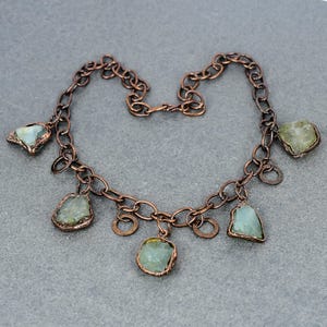 May include: A necklace with a copper-coloured chain and irregular links. It features light green, irregularly shaped gemstones as pendants. Small, round copper accents are also included, enhancing the necklace's distinctive appearance.