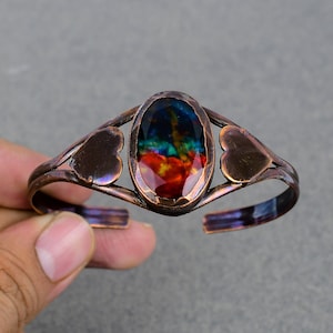 May include: A copper cuff bracelet featuring an oval-shaped, multi-colored stone. The stone displays a vibrant array of colors, including red, blue, and green. The bracelet has heart-shaped accents on each side of the stone.