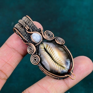 May include: A wire-wrapped pendant featuring a central cowrie shell with a smooth, cream-colored surface and dark edges. The shell is encased in dark, oxidized copper wire with intricate spiral details and a small, round, white moonstone accent.